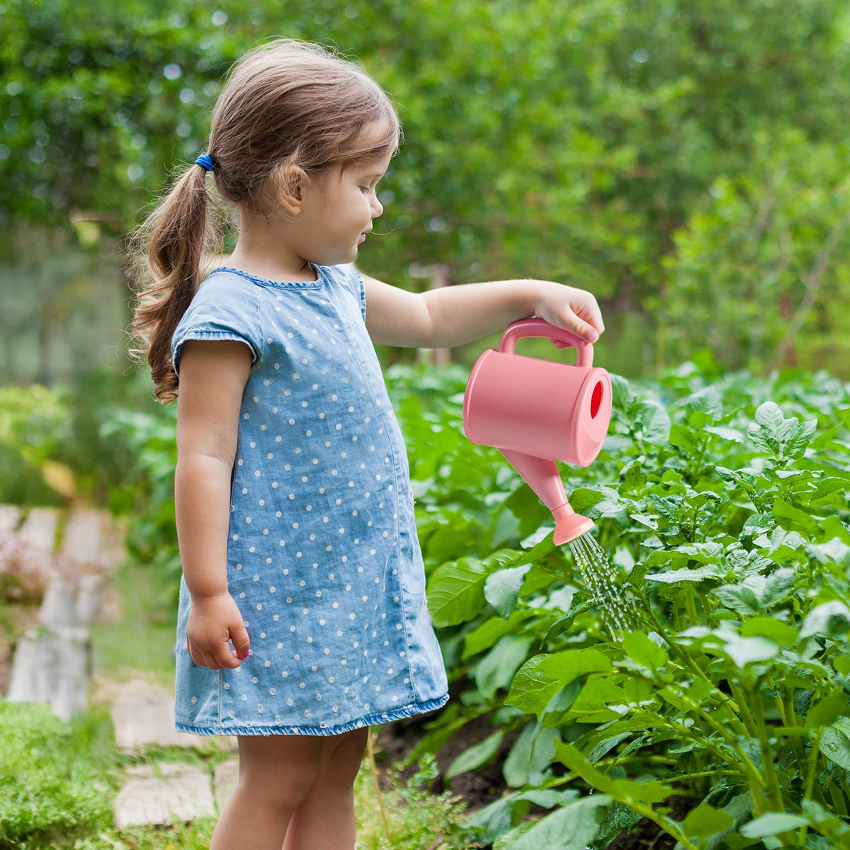 Young girl in blue dress watering garden plants with ROBUD Kids pink watering can in a lush green outdoor garden bed