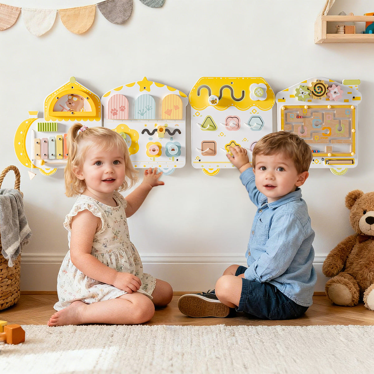 Two young children sit on a wood floor in front of the ROBUD yellow spring train busy board mounted at low toddler height.