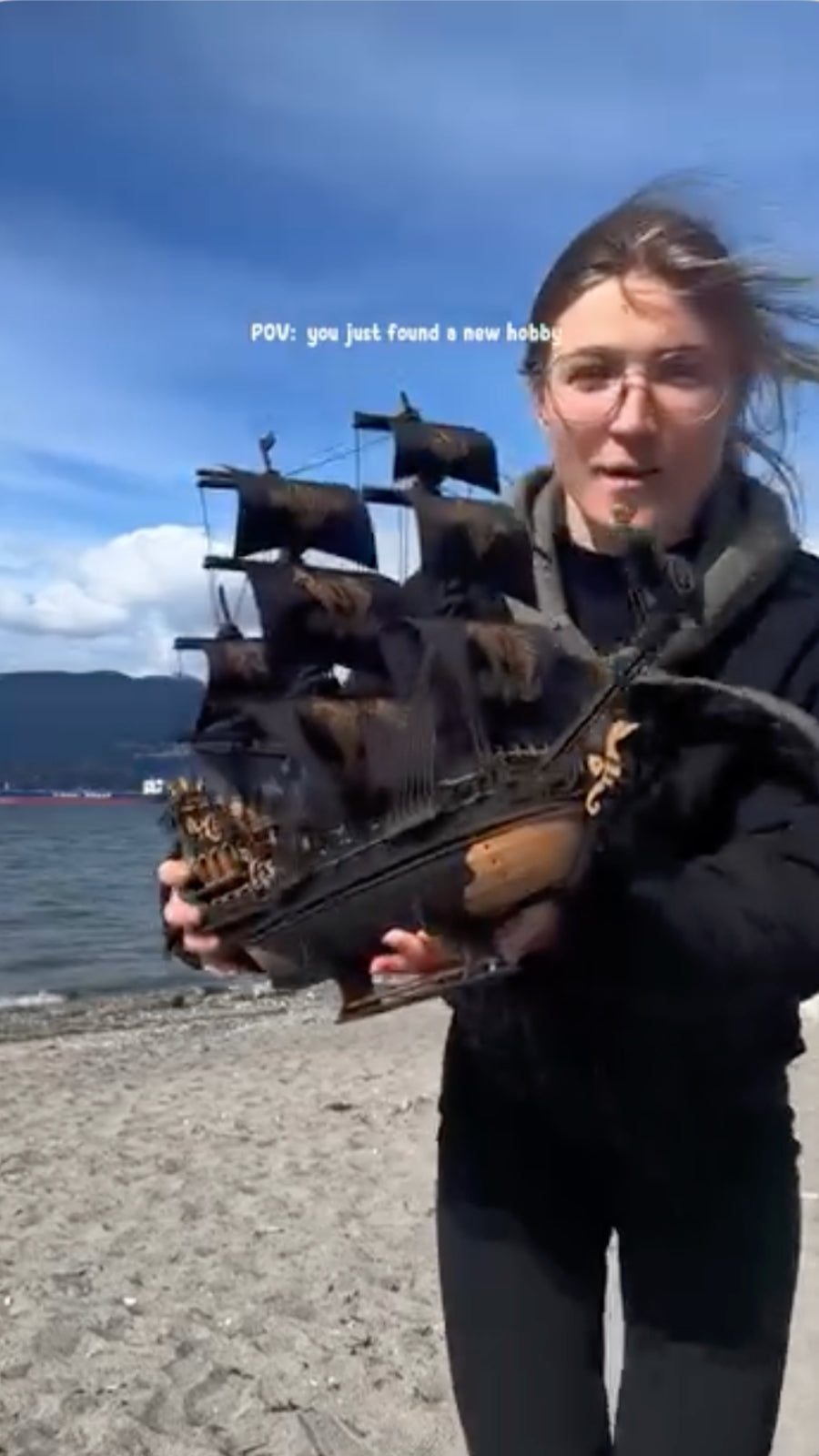 Man displaying a dark wooden pirate ship model outdoors beside water and mountains
