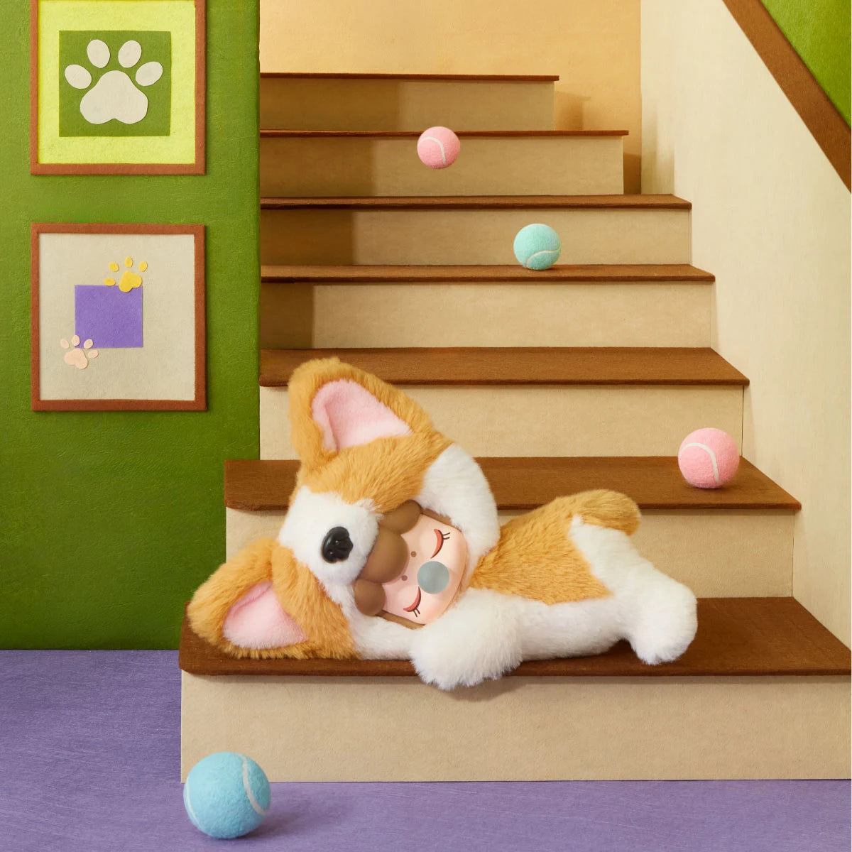 Plüsch-Corgi mit Puppengesicht und Blase liegt auf einer Miniatur-Holztreppe, umgeben von rosa und blauen Tennisbällen, Wandbildern mit Pfotenabdrücken und grünen Wänden.
