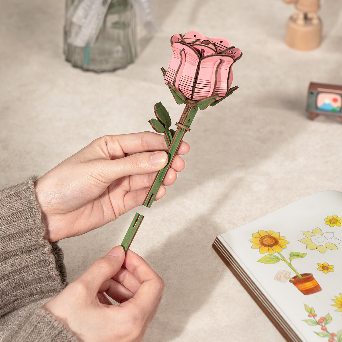 Der Stiel der rosa Rowood Holzrose wird von einer Hand über eine Bastelszene auf dem Tisch gehalten.