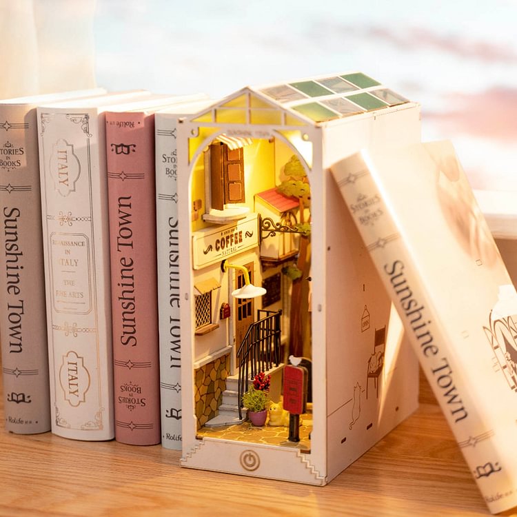 Rolife Sunshine Town book nook glowing between real books on a shelf with a lit cobblestone alley and warm window light