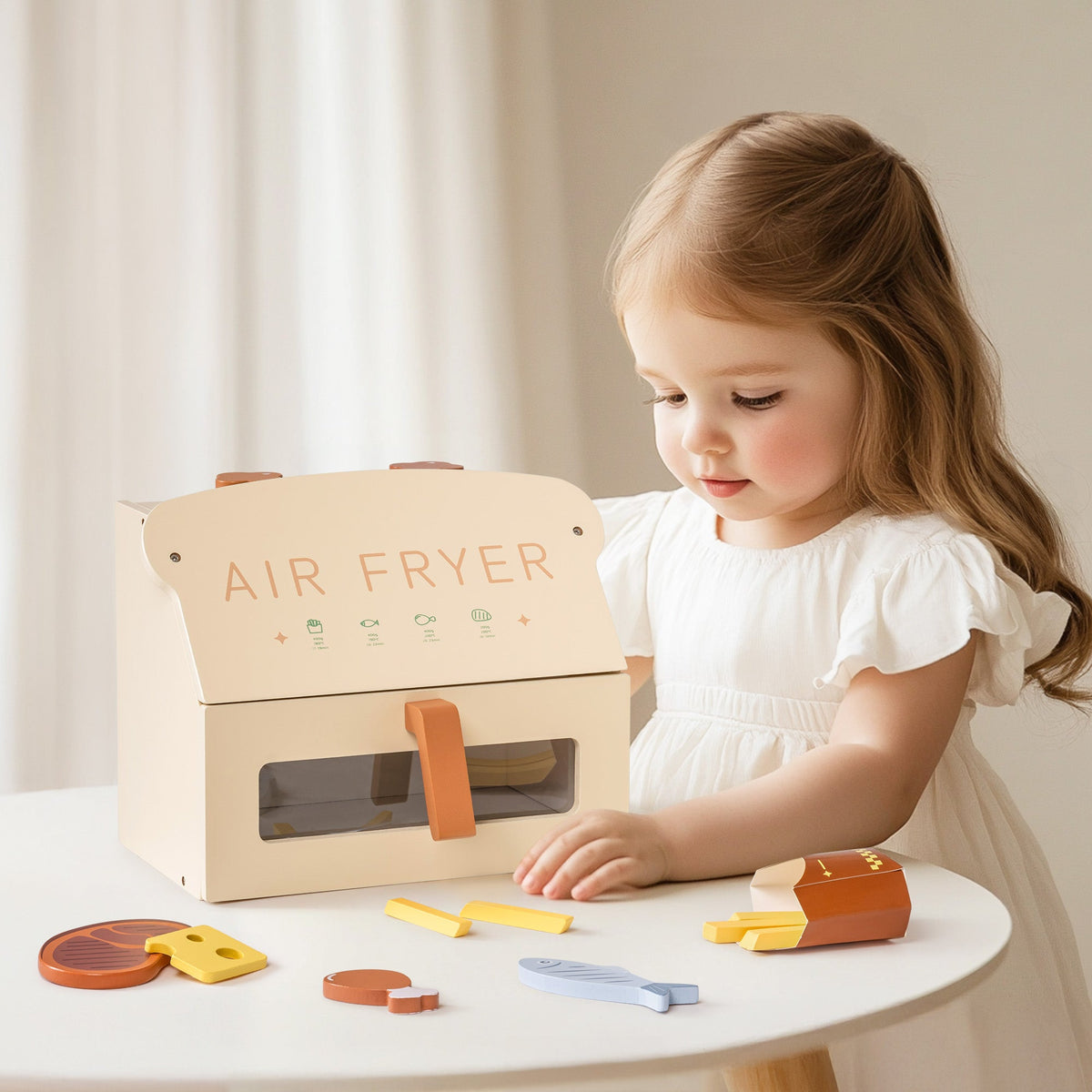 Young girl in a white dress looking at the ROBUD creamy wooden play air fryer on a round table with fish, steak, cheese, fries, and chicken leg pieces nearby