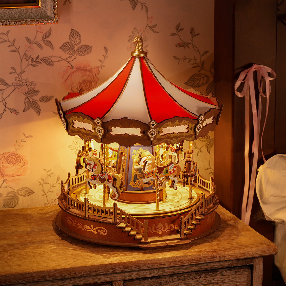 Rolife Classic Carousel AMC01 placed on wooden dresser beside a pink armchair with warm bokeh fairground lights behind