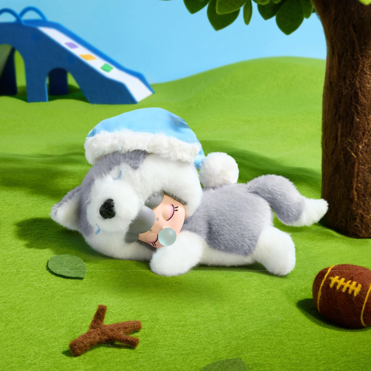 Grau-weißer Plüsch-Husky mit Puppengesicht liegt auf grünem Filzrasen neben einer blauen Rutsche, einem Baum, einem Blatt, einem Zweig und einem braunen Football.