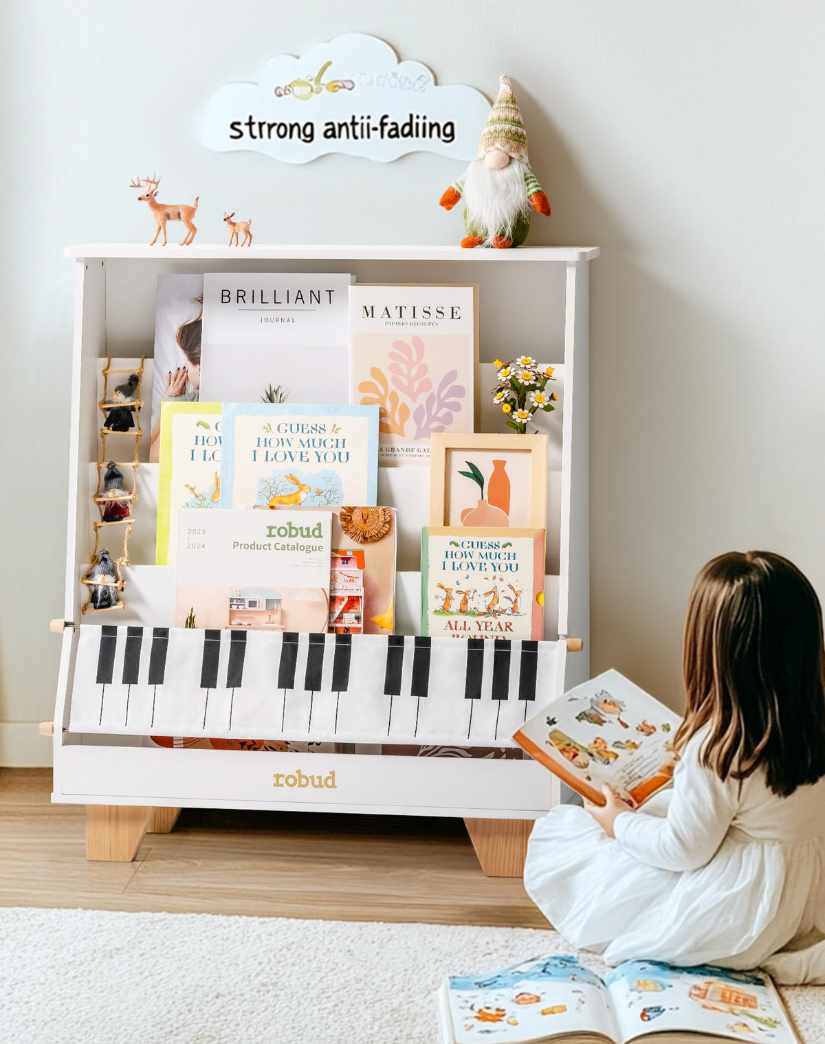 A young girl in white sitting on a rug reading a book beside the ROBUD Wooden Kids Bookshelf Piano white version stocked with picture books and an art print in a bright home setting
