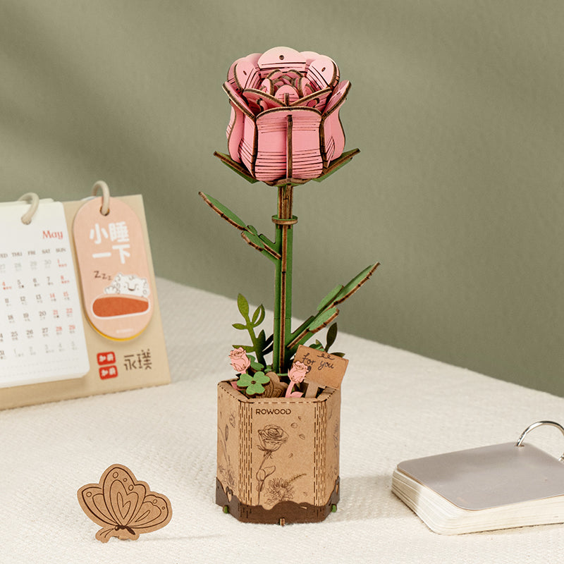 Die rosa Rowood Holzrose steht in einem Sockel in Korkoptik, daneben die Verpackung und kleine Schreibtisch-Accessoires.