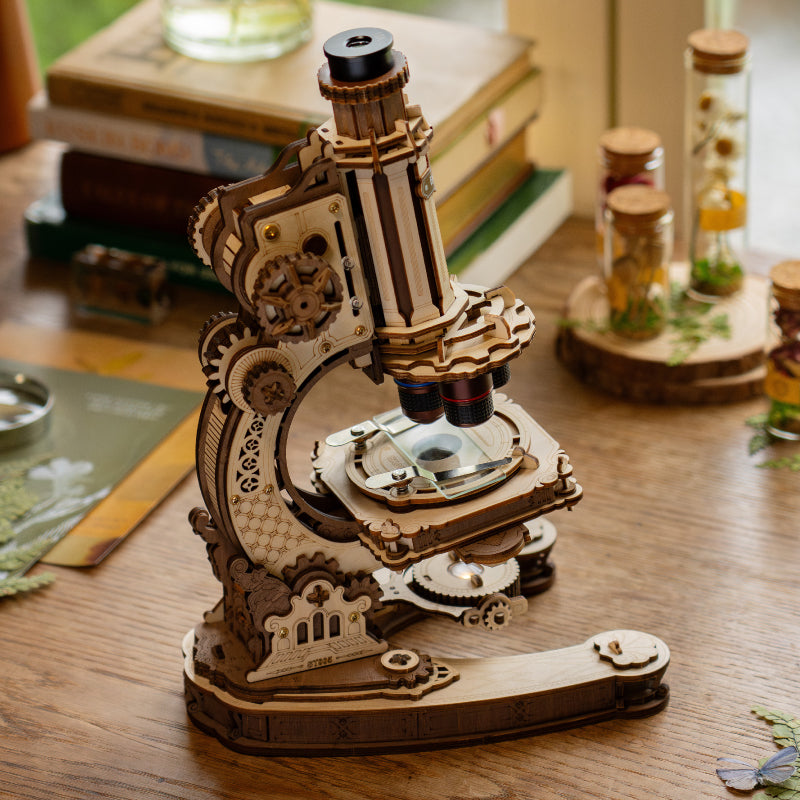 ROKR wooden microscope model kit with brass toned gears displayed on a table beside glass specimen bottles and dried flowers