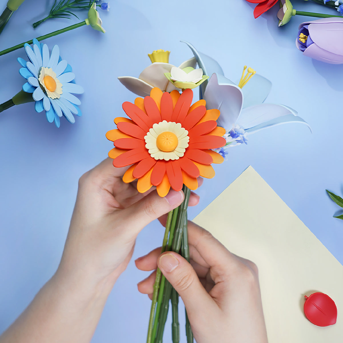 Hands holding a Rowood foam orange gerbera stem on bright blue background beside a small watering can