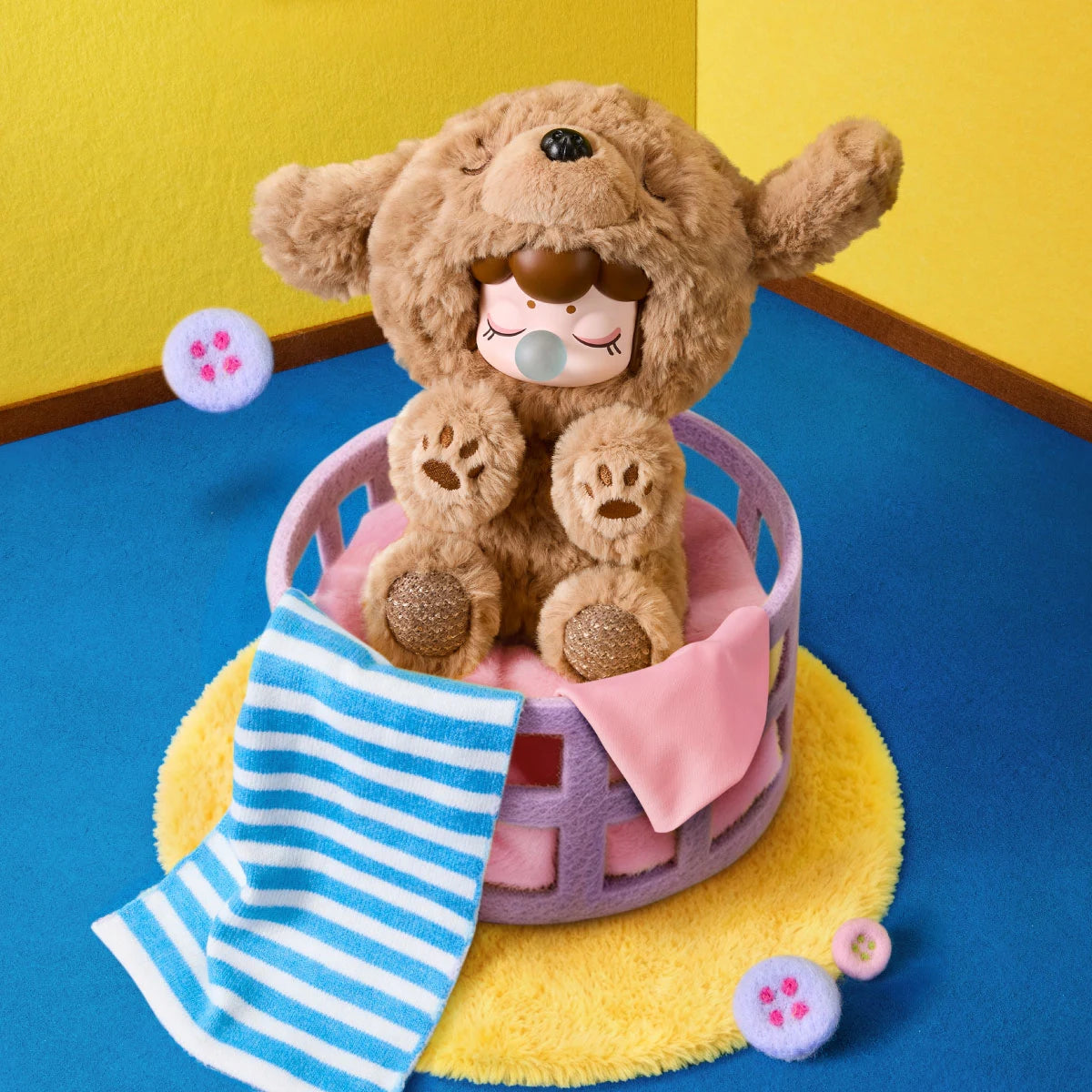 Braune Plüschfigur mit Welpenkapuze, geschlossenen Augen und blauer Blase sitzt in einem lavendelfarbenen Korb auf einem gelben Teppich, mit gestreiftem Handtuch und rosa Tuch.