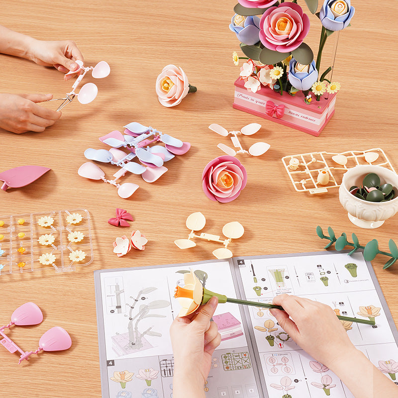 Rowood Infinite Smiles AD011 floral art kit assembly, hands assembling pink and blue plastic flower petals with instruction booklet and bouquet on a wooden table
