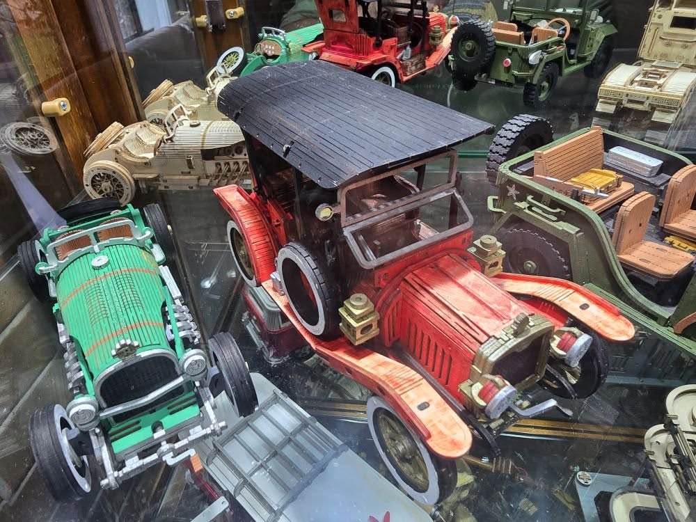 Customer-built ROKR wooden model car collection displayed in a glass case featuring red vintage sedan, green racing car and other hand-painted vehicles