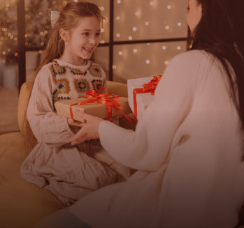 Gifts for kids lifestyle scene showing a girl receiving a red-ribboned gift from an adult in a warm Christmas setting
