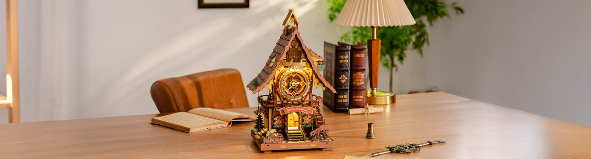 Robotime Online Wonderland themed banner showing a glowing ROKR wooden cuckoo clock house with intricate gear and floral motifs beside stacked books and a table lamp on a wooden desk.