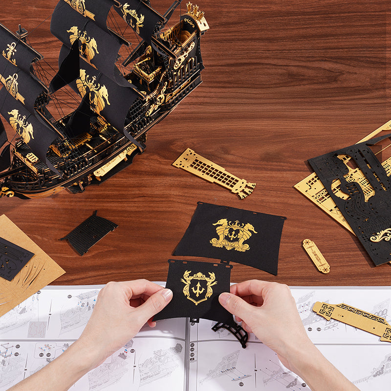 ROKR Seahorse Barque MCB02 assembly view, hands assembling black sail panel with gold crest, wooden ship and parts spread on table