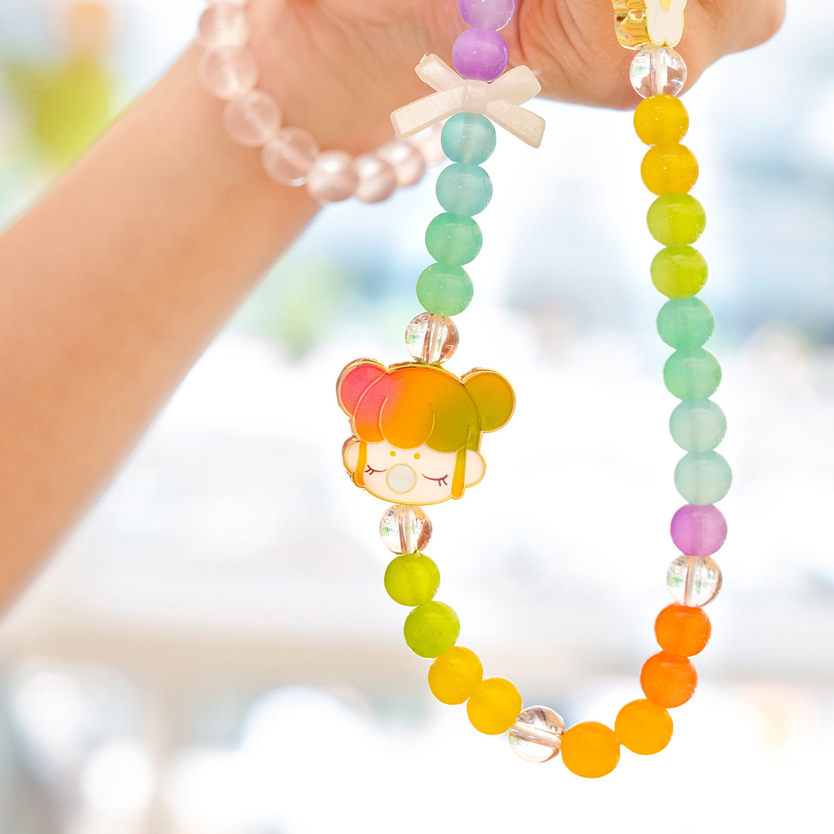 Hand holding a Rolife Nanci Gift Bag rainbow bead lanyard close up, showing the multicolor enamel character charm against a blurred outdoor background.