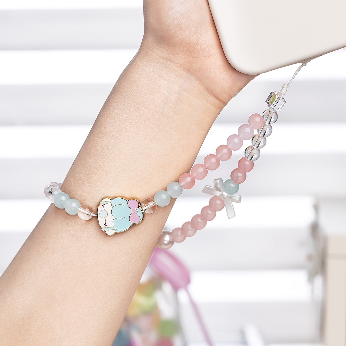 Wrist wearing a Rolife Nanci Sweet Time bead bracelet with a blue bunny charm while holding a phone with a pink bead lanyard, indoor lifestyle shot.