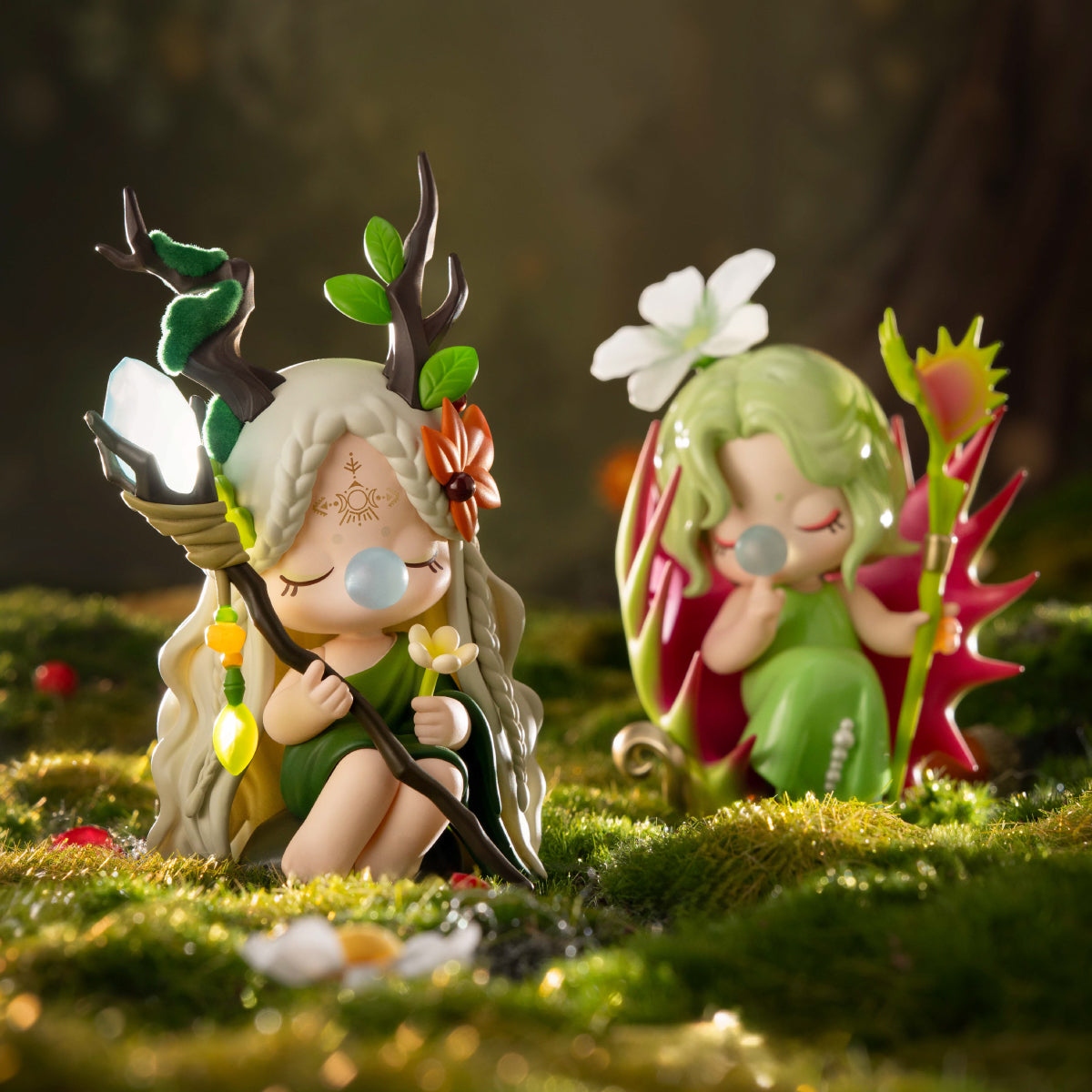 Two fairy-themed figurines with floral headdresses seated together in a mossy forest scene