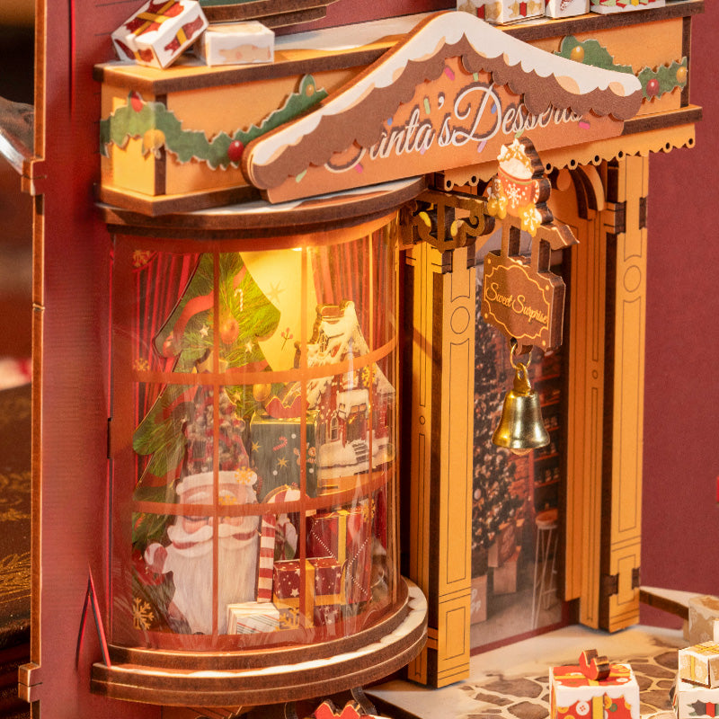 Close-up of Rolife Christmas book nook showing Santa's Desserts shop bay window with illuminated Santa figurine and a gold bell sign.