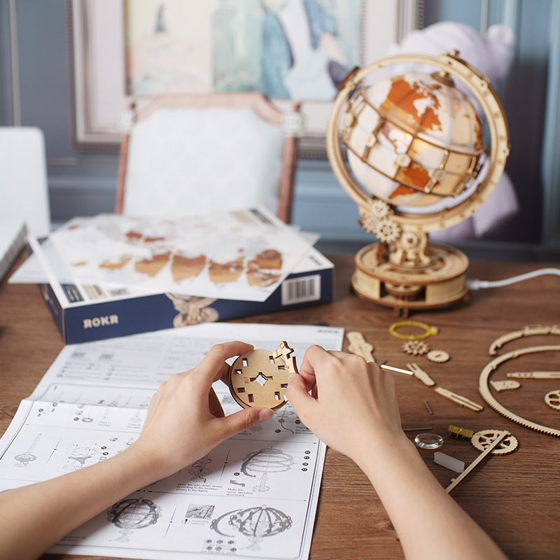 ROKR Luminous Globe ST003 assembly scene, hands fitting wooden sphere piece with instruction booklet and puzzle parts spread on desk