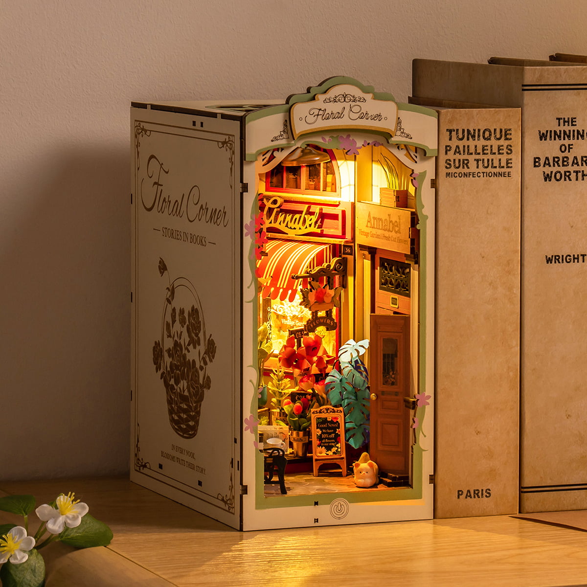Rolife Floral Corner TGB09 Book Nook displayed between real books on wooden shelf showing illuminated flower shop with warm ambient lighting