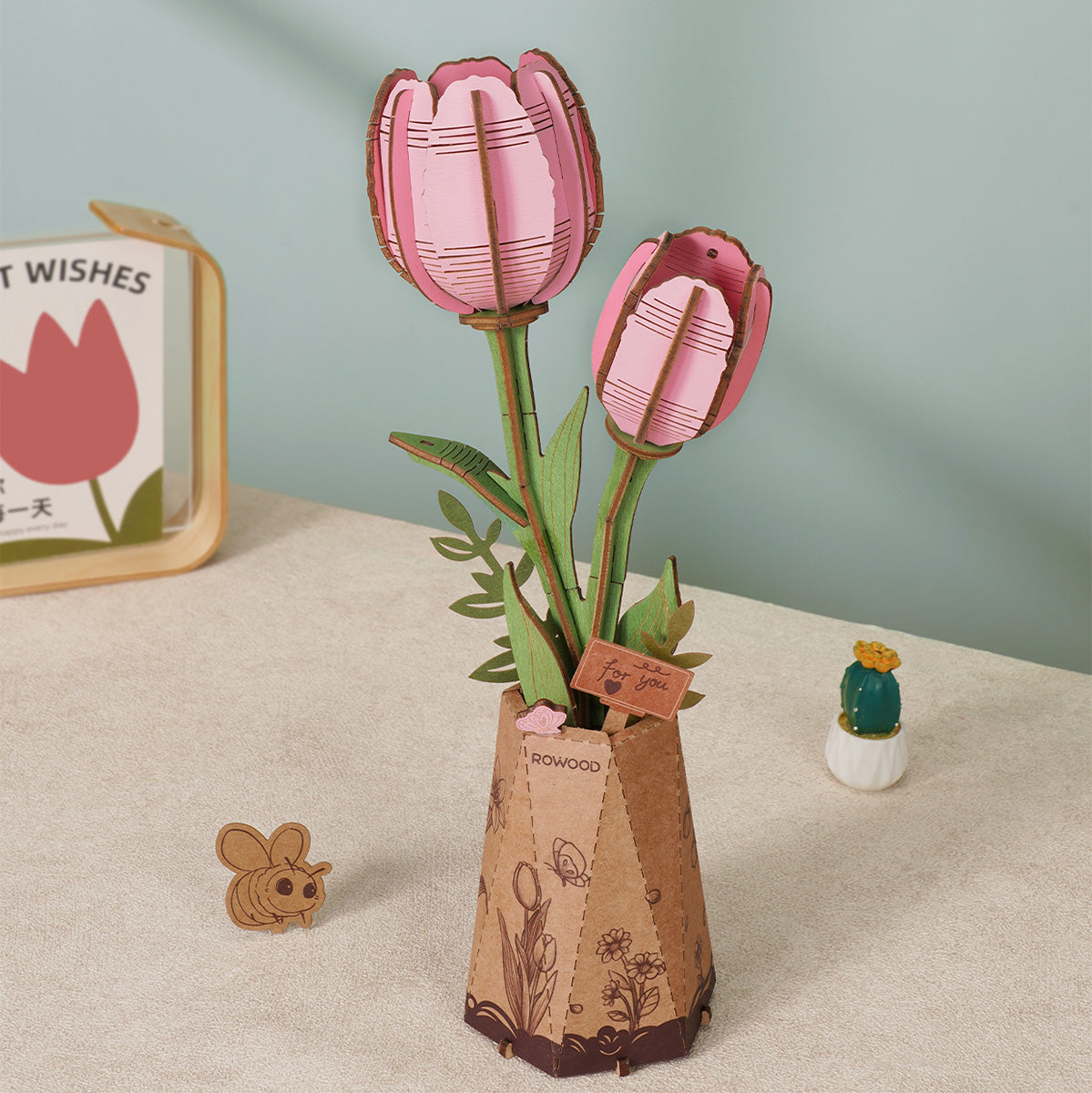 Rowood Holzblumenstrauß-Bausatz Lila Orchidee TW091, zusammengebaut mit mehreren Blüten und grünem Blattwerk, in einer Kraftpapier-Vase auf einem Schreibtisch neben einem Kaktus