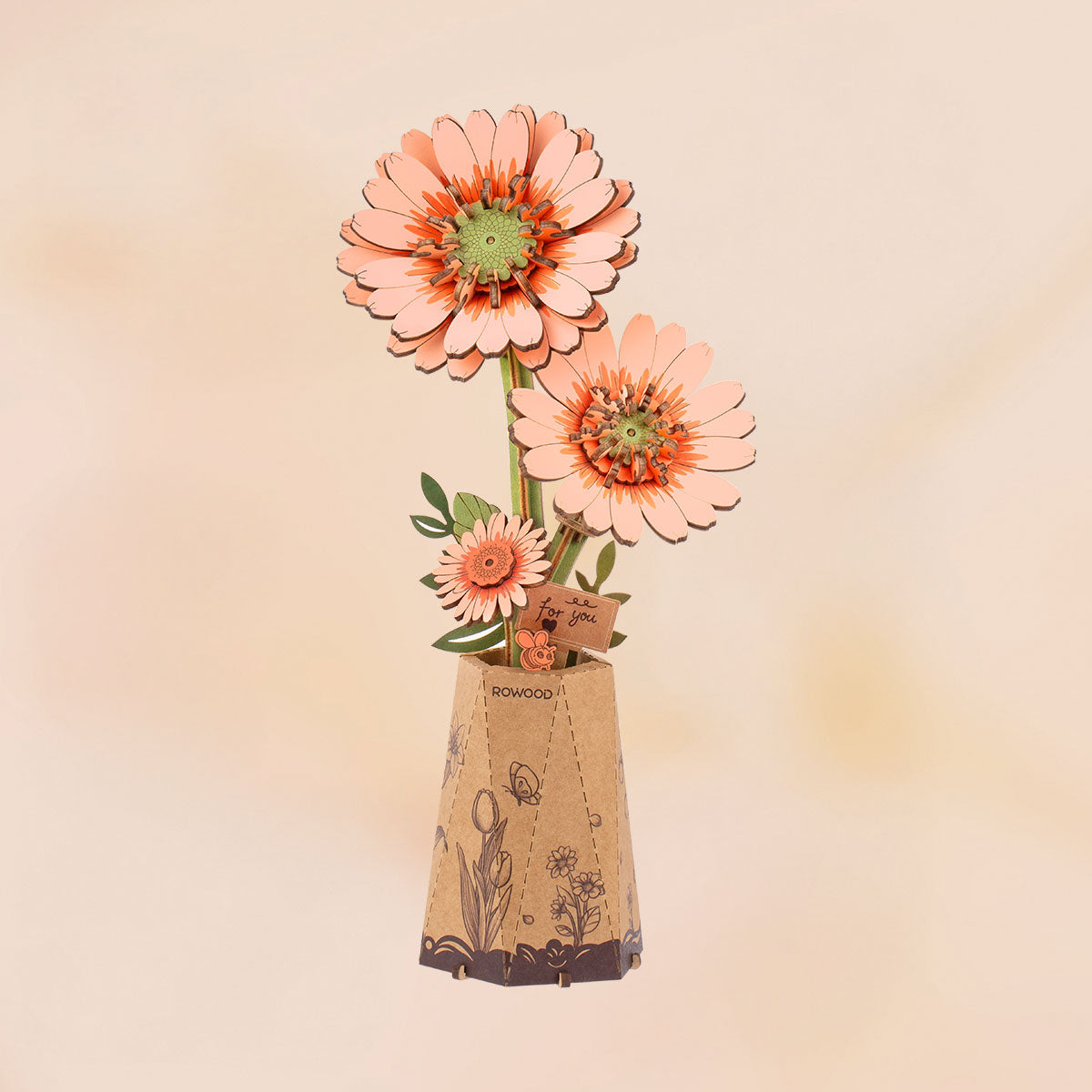 Rowood DIY-Holzblumenstrauß TW111: fertiges Gerbera-Modell mit zwei großen Blüten in Pfirsich und Koralle und einer kleineren Knospe in einem Kraftpapier-Topf vor pfirsichfarbenem Hintergrund.