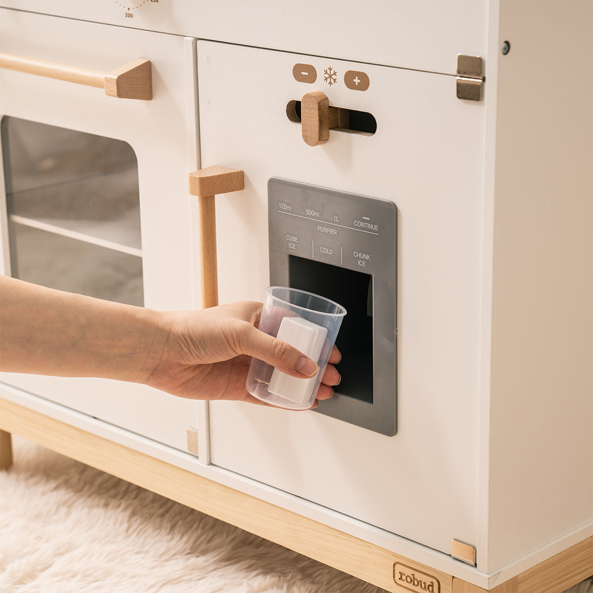 Hand pressing cup under ROBUD white wooden play kitchen ice and water dispenser on right cabinet.