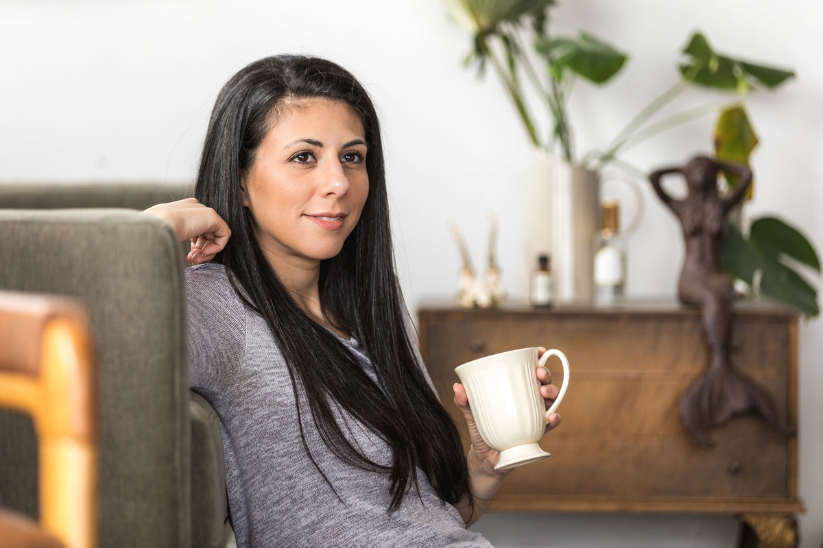 Young woman with long dark hair relaxing on grey sofa holding white coffee mug in cozy home living room lifestyle setting