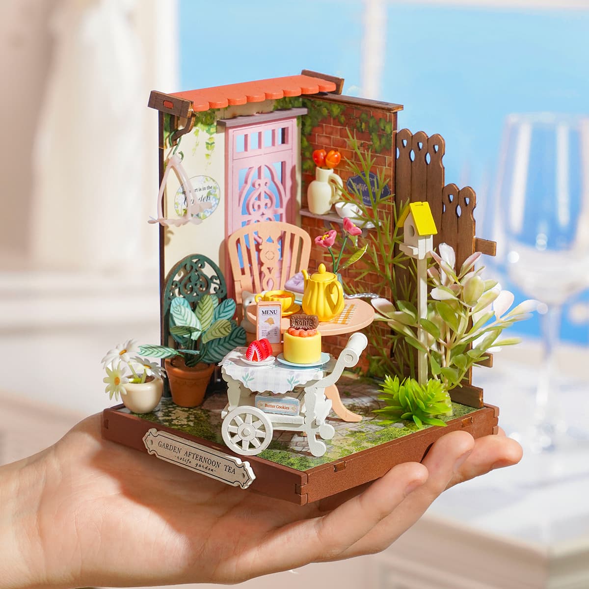 Rolife Fancy Tea Yard miniature garden scene held in a hand showing flower wall, pink chair and cake cart