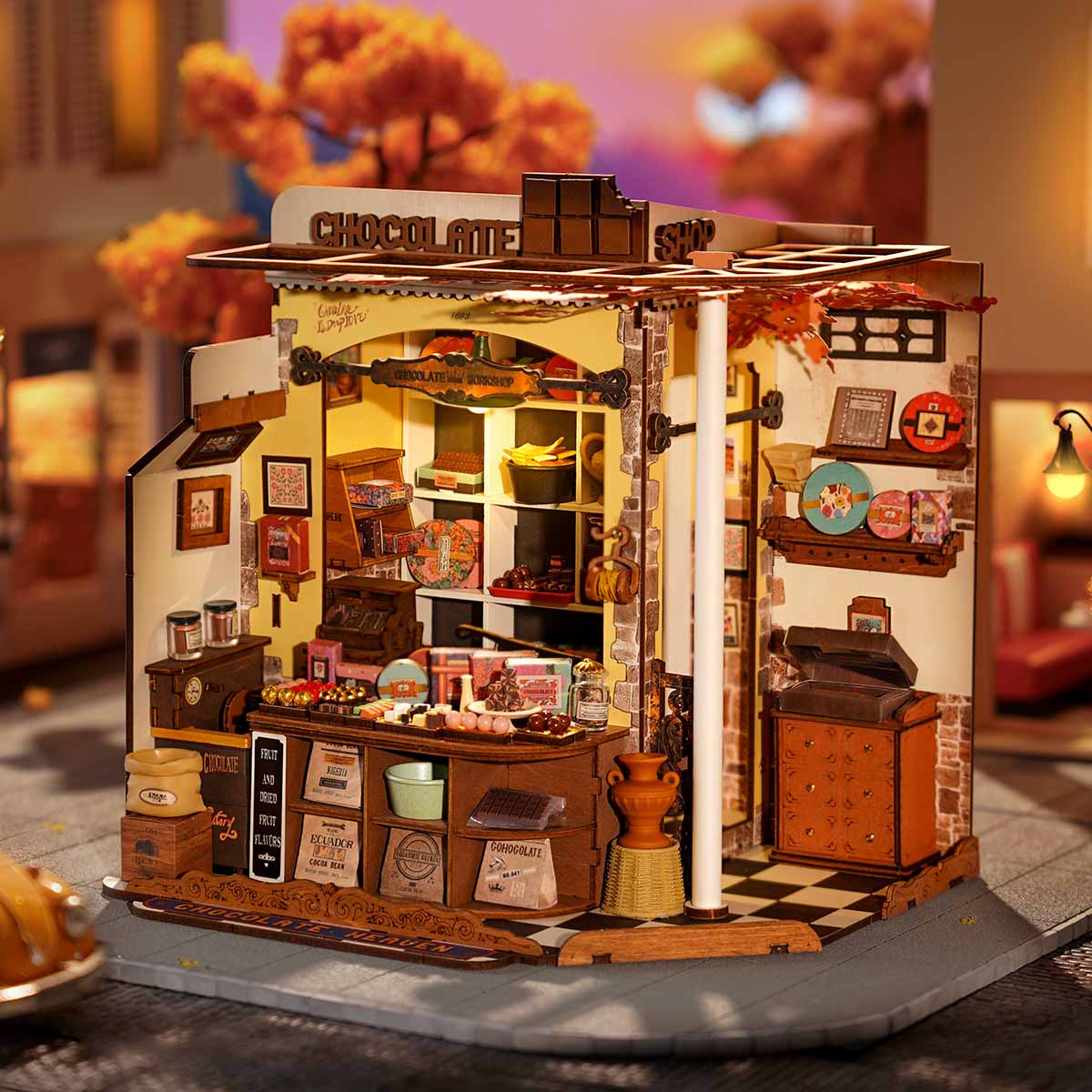 Rolife Henry's Chocolate DIY miniature house DG174 lifestyle side view showing the assembled illuminated shop with autumn leaf decorations and a wooden rooftop chocolate bar display outdoors.