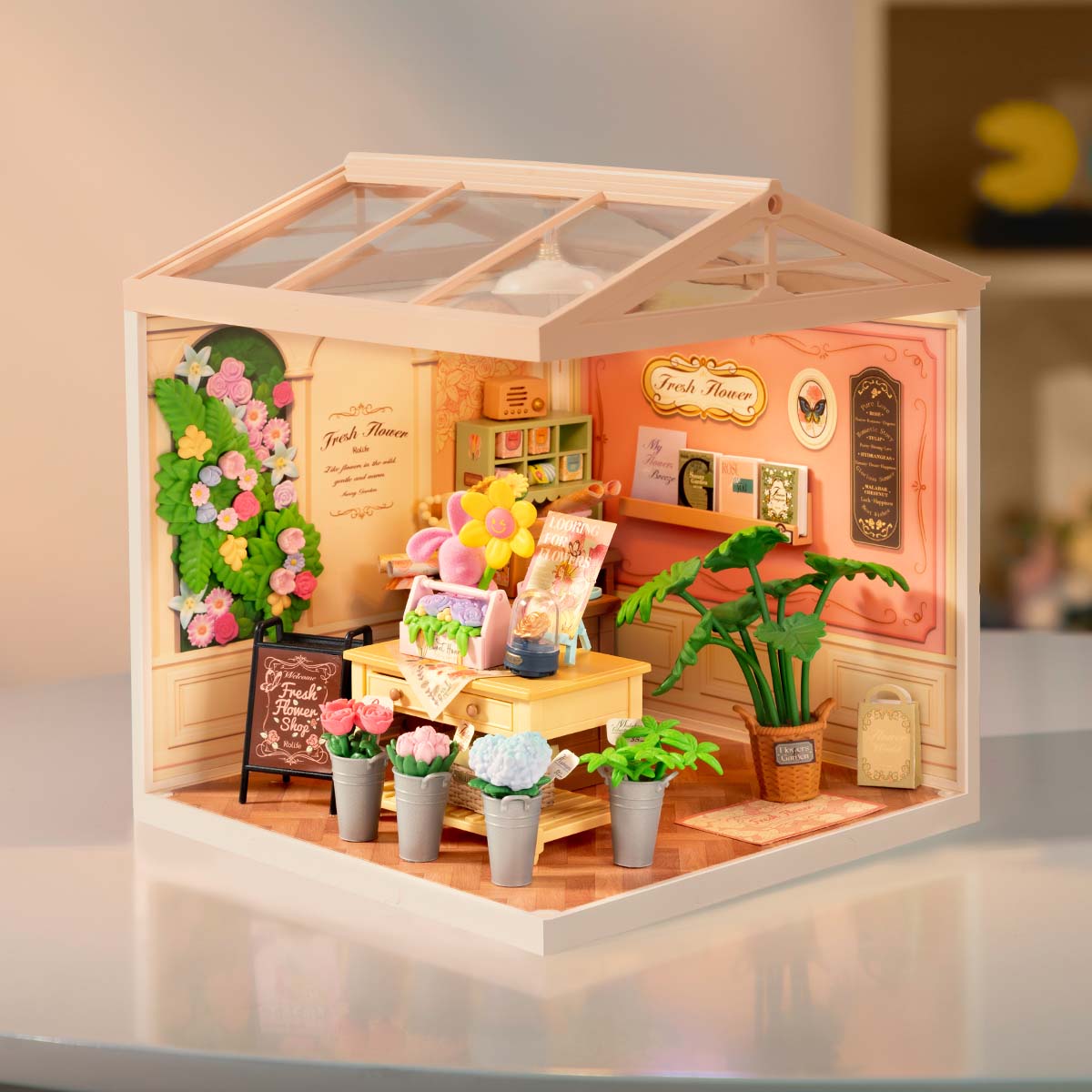 Rolife Fresh Flower Shop DIY miniature house DW017B lifestyle scene showing the illuminated flower shop with colorful blooms along the archway, a central work table, and tropical potted plants.