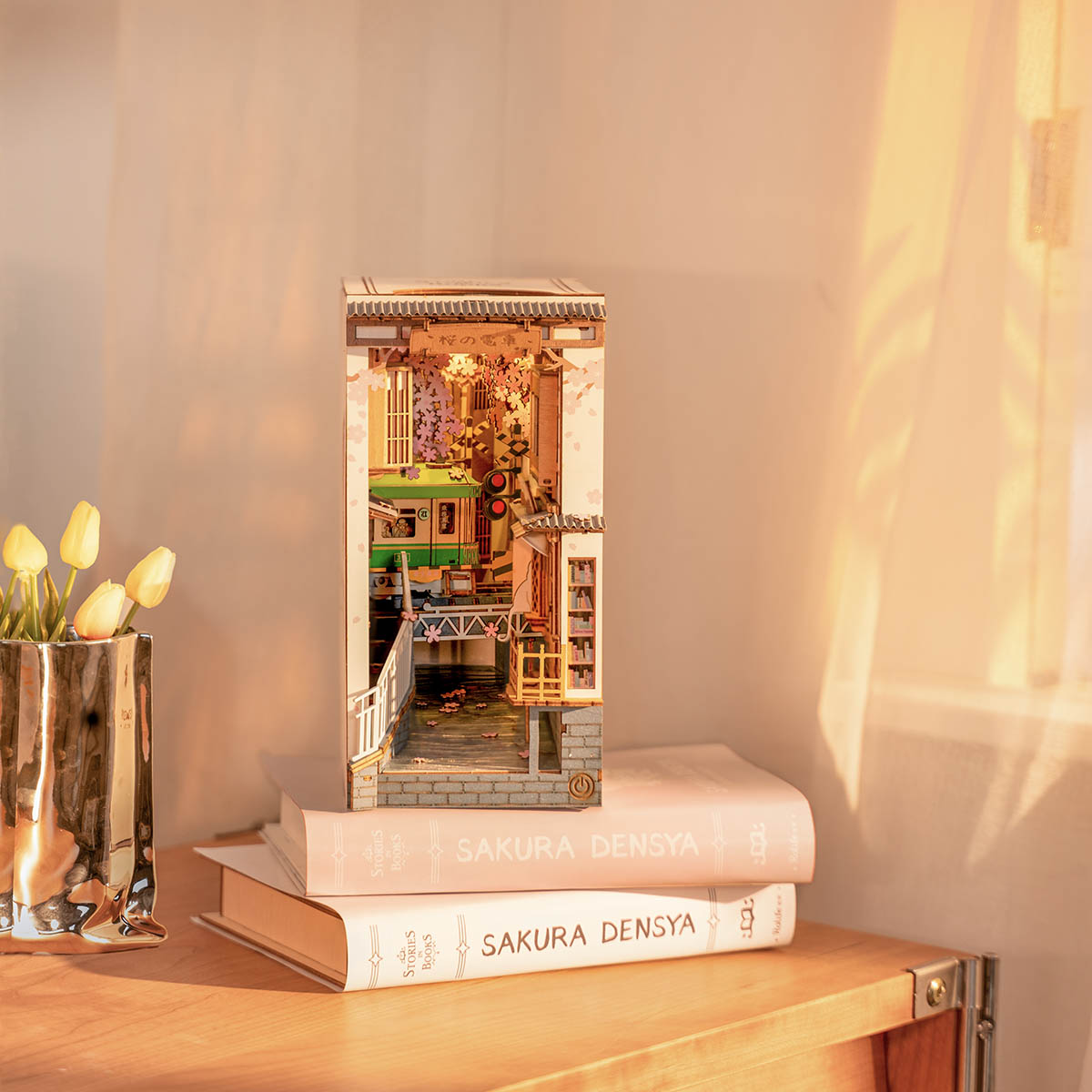 Rolife Sakura Densya book nook displayed on stacked books beside tulips in warm afternoon light