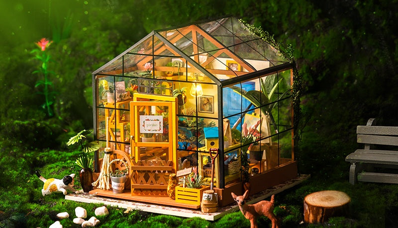 Rolife Miller's Garden DIY miniature house model with glass greenhouse walls and warm interior lighting displayed outdoors on green mossy ground with a deer figurine and garden bench nearby