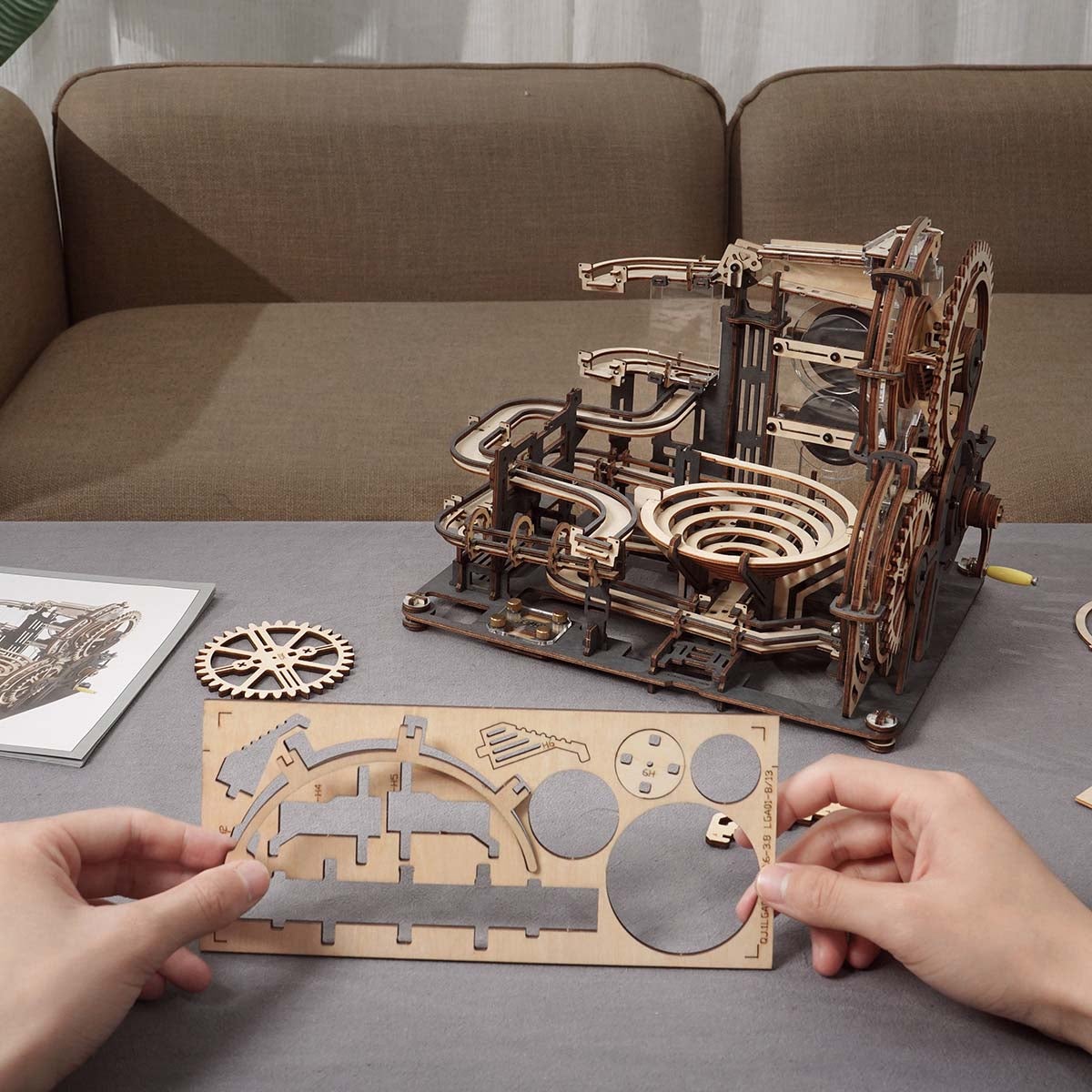 Hands holding a laser-cut wooden gear sheet beside the partially assembled ROKR Marble Night City run on a grey table