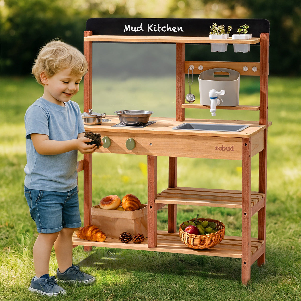 Toddler boy playing at the ROBUD outdoor wooden mud kitchen in a backyard garden, holding stainless cookware beside the sink and stove unit
