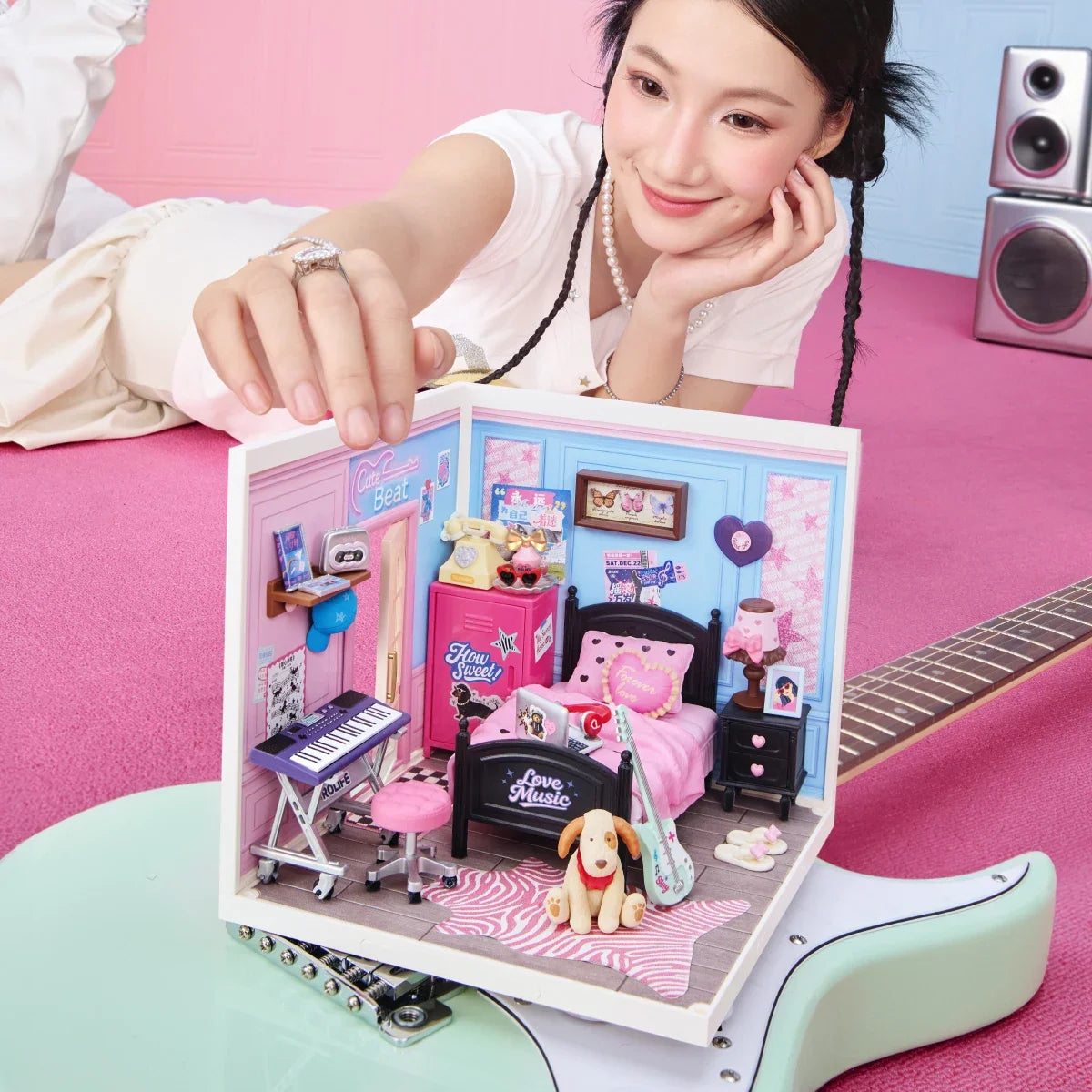 Rolife-Miniatur-Schlafzimmer auf einer rosa Fläche, mit einer Hand neben dem Modell und einem sichtbaren Gitarrenhals im Vordergrund.