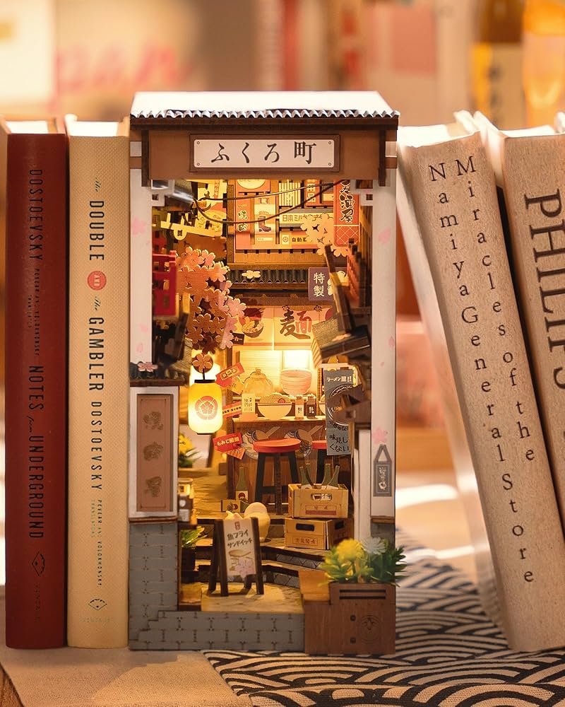 Rolife Sakura Wine Alley TGB11 book nook standing between real books on a bookshelf showing the warm amber street scene with cherry blossoms