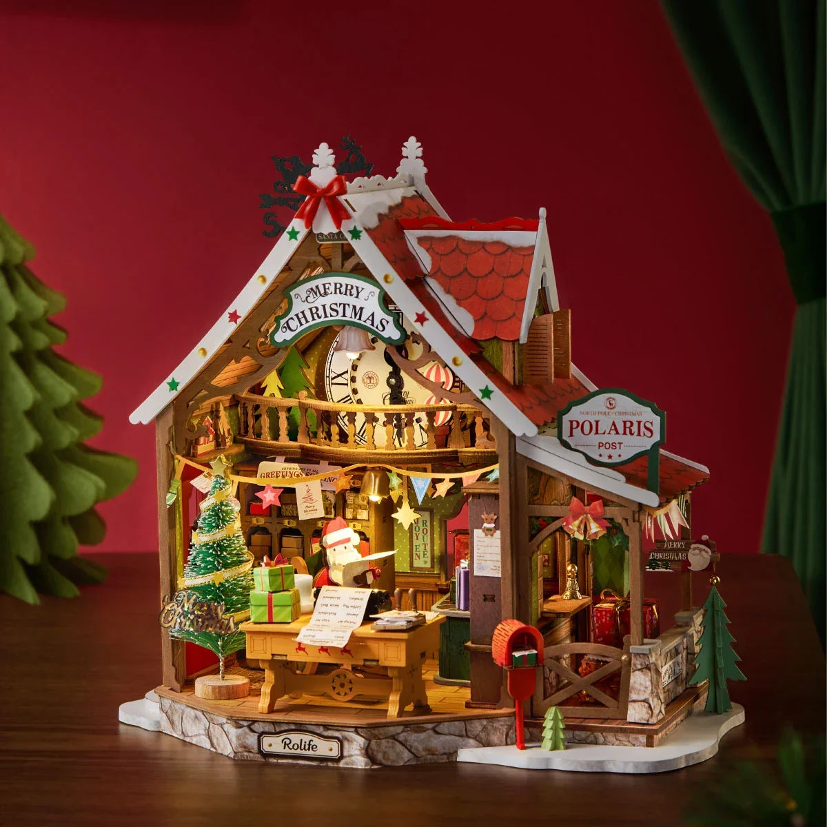 Hölzernes Rolife-Weihnachtspostamt im Miniaturformat mit rotem Dach, Weihnachtsmann am Schreibtisch, geschmücktem Baum, rotem Briefkasten und Polaris Post Schild, warm beleuchtet vor rotem Hintergrund.