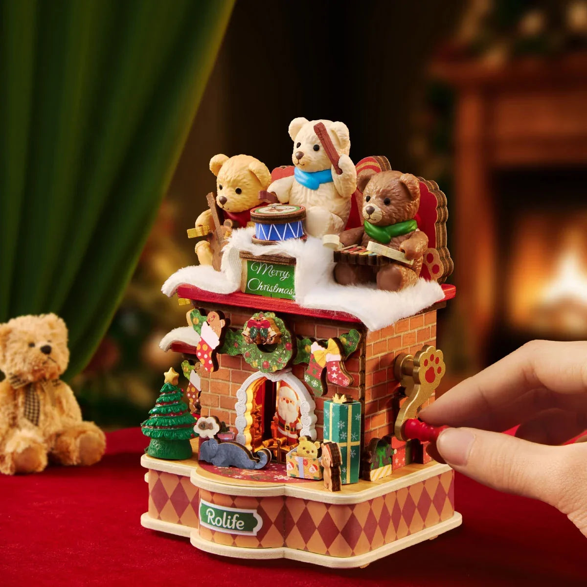 Eine Hand dreht den roten Kurbelgriff an der Seite der Rolife-Weihnachts-Kaminspieluhr, mit Teddybären oben und einem Plüschbären daneben auf rotem Stoff.