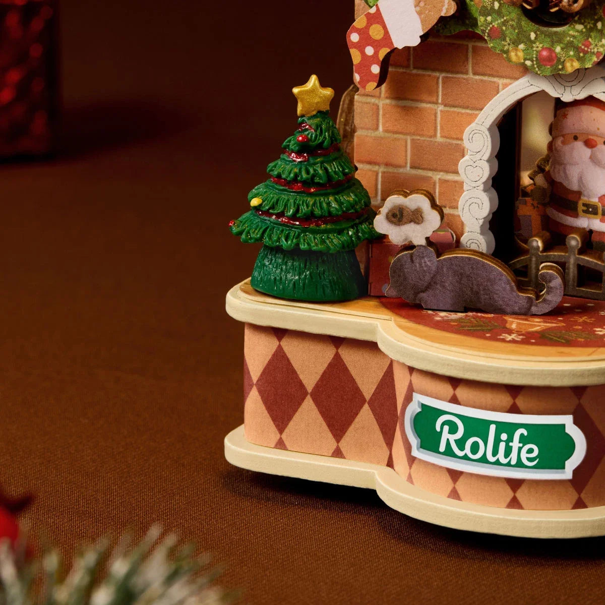 Nahaufnahme der Rolife-Modellbasis mit grünem Rolife-Schild, Seiten im Rautenmuster, Miniaturweihnachtsbaum mit Stern, grauer Katze und Weihnachtsmannfigur im Kamin.