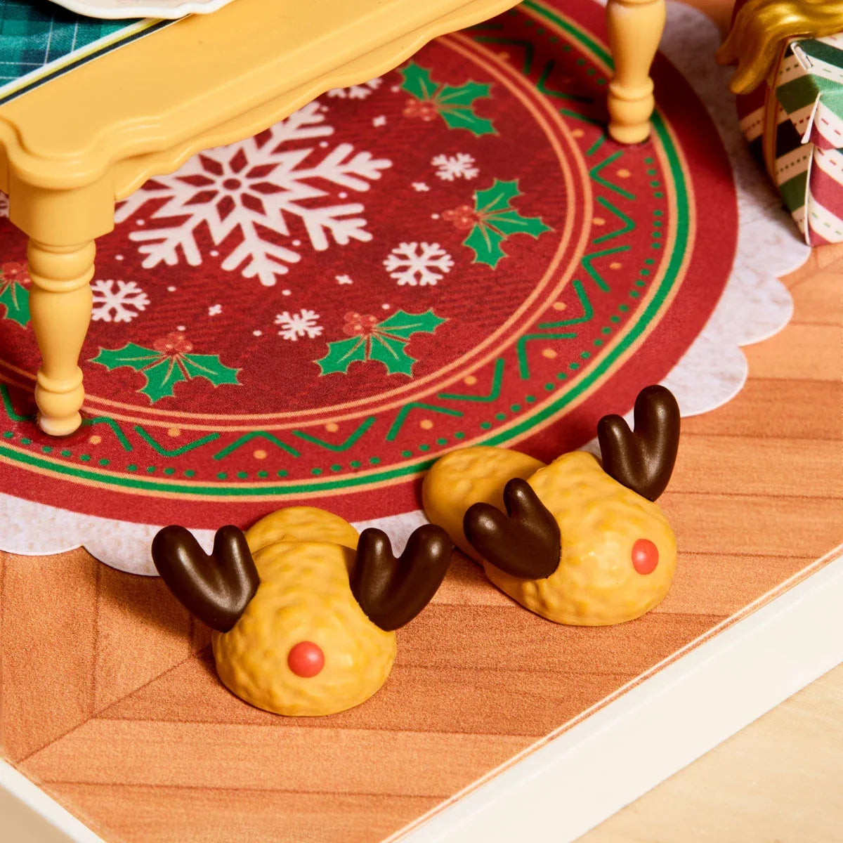Nahaufnahme von zwei Miniatur-Rentierpantoffeln mit braunem Geweih und roten Nasen auf einem Teppich mit Schneeflockenmuster.