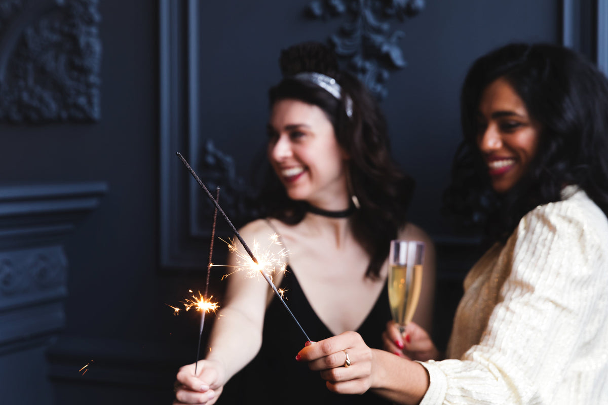 Two women smiling and celebrating with glowing sparklers and champagne glasses against dark moody background for a festive